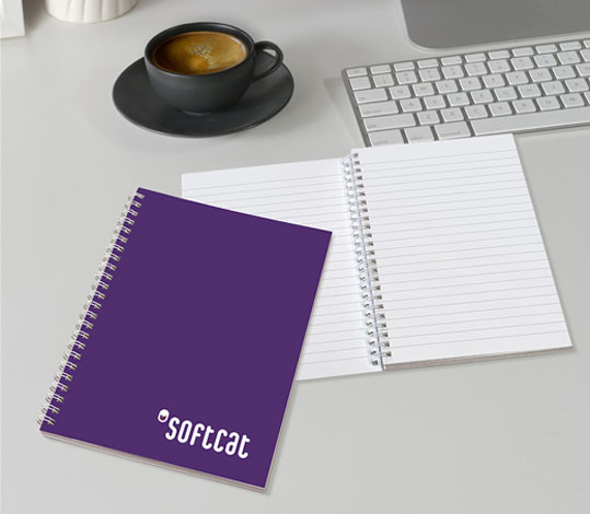 Softcat-branded purple spiral notebook placed on a desk next to an open lined notebook, a keyboard, and a cup of coffee.